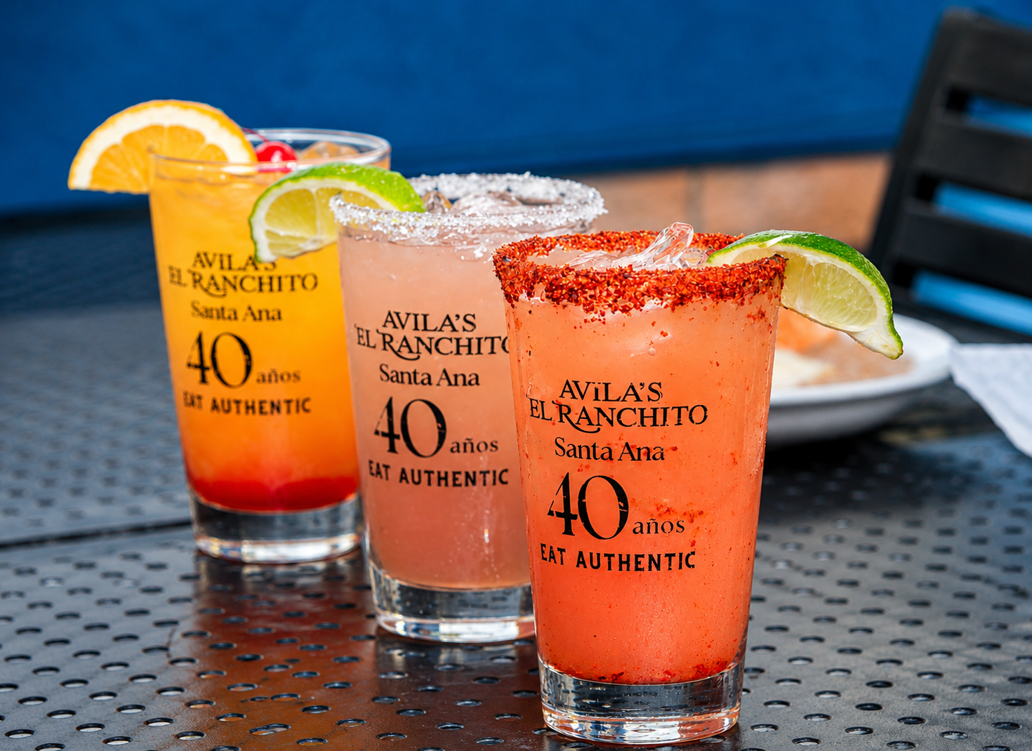 Three branded Avila's El Ranchito 40 años glasses filled with colorful margaritas on a patio table