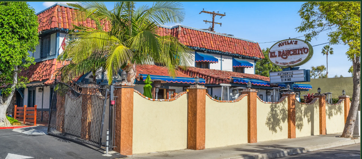 Avila's El Ranchito Santa Ana exterior on a sunny day: white stucco walls, red clay tile roof, blue awnings and the restaurant's marquee sign behind a cream wall with palm trees